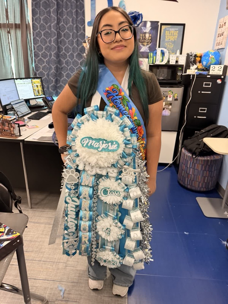 Woman with turquoise hair wearing a sash with the word "Majorette" on it in a classroom.