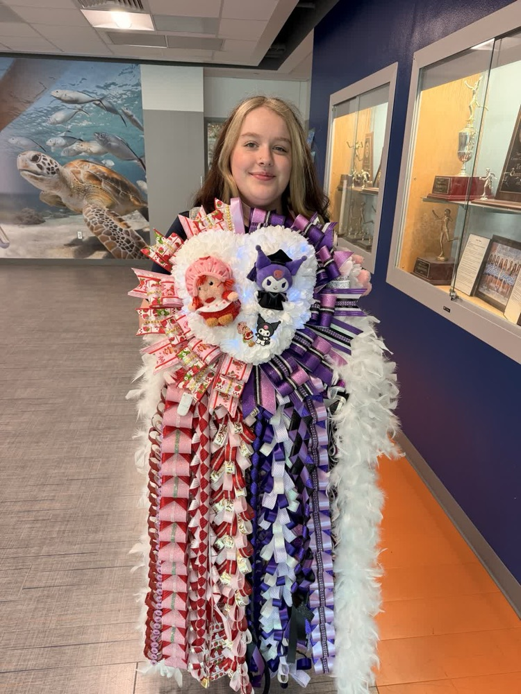 Girl in a hallway, wearing a colorful, decorative sash with a large, round center piece.