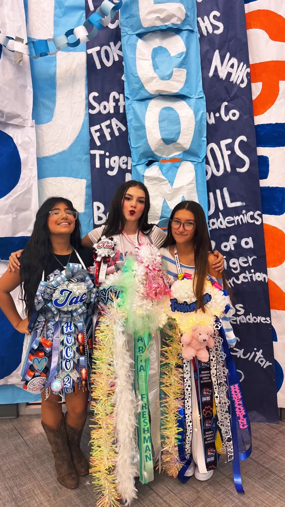 Three girls in festive attire stand together in front of a blue and orange banner. Each holds decorative items.