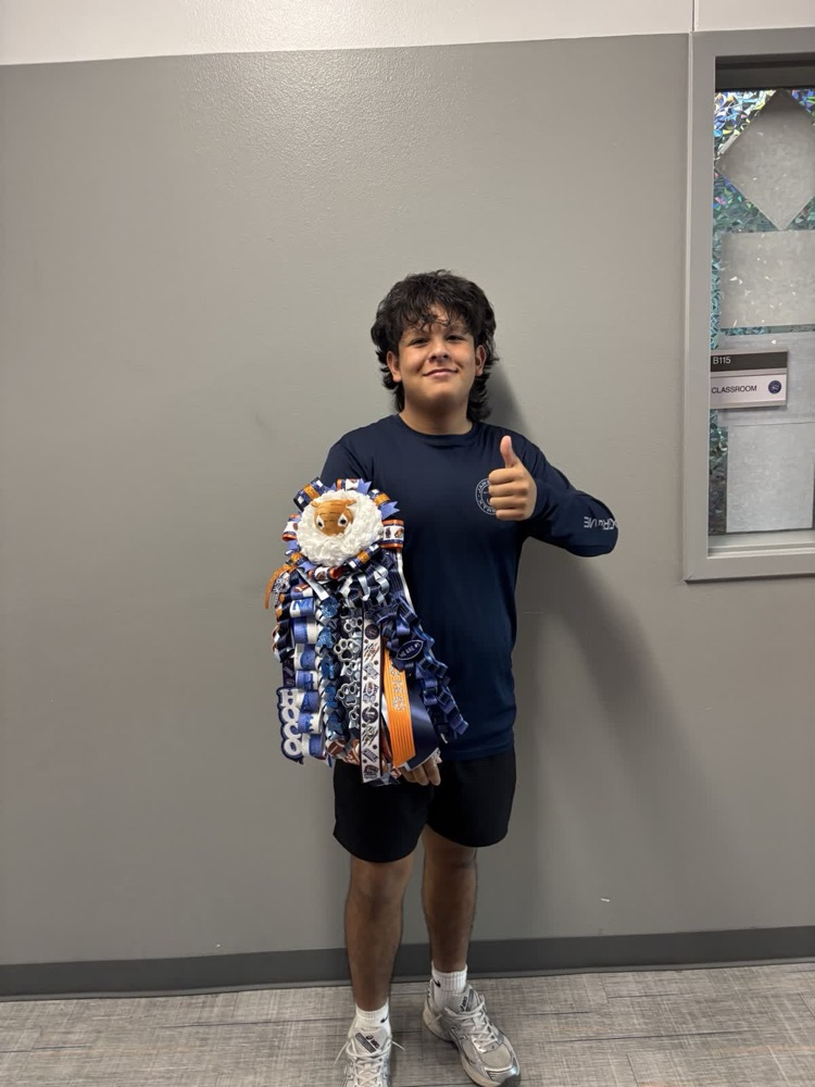 Boy stands, smiling, with a blue ribbon and pom-pom, giving a thumbs-up. He wears navy shirt, black shorts, white shoes. Behind him, gray wall and glass window.