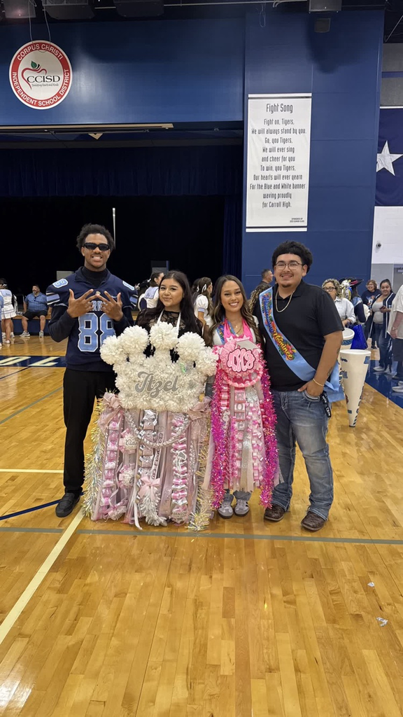 Four students in a gym with a banner on the wall. One holds a pom-pom-covered table.