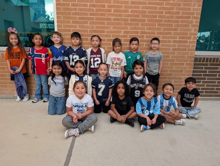 Children in sports jerseys sit and stand in two rows outside a brick building.