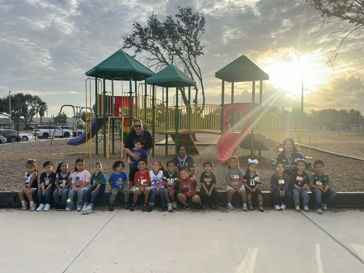 Group of children and adults sit and stand near a playground at sunset. Playground has a slide, climbing structure, and swings.