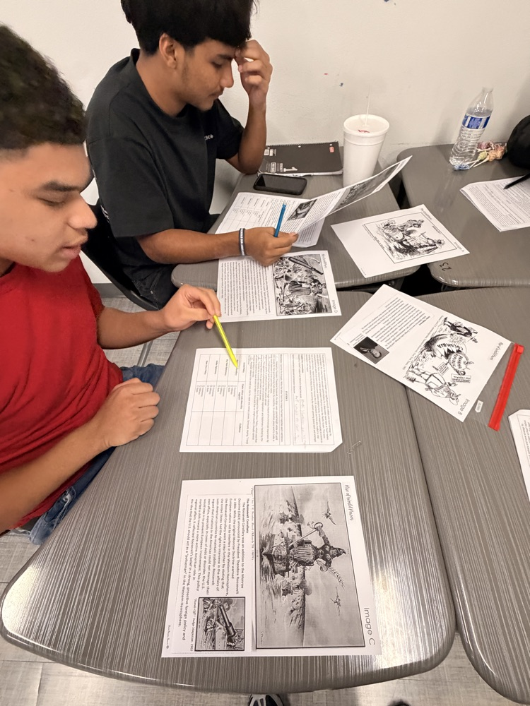 Two students sit at a table with papers and a pencil. One holds a pencil while the other looks at a paper.
