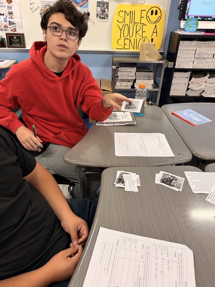 Two students seated at tables, one pointing at a paper with a pen, both surrounded by papers.