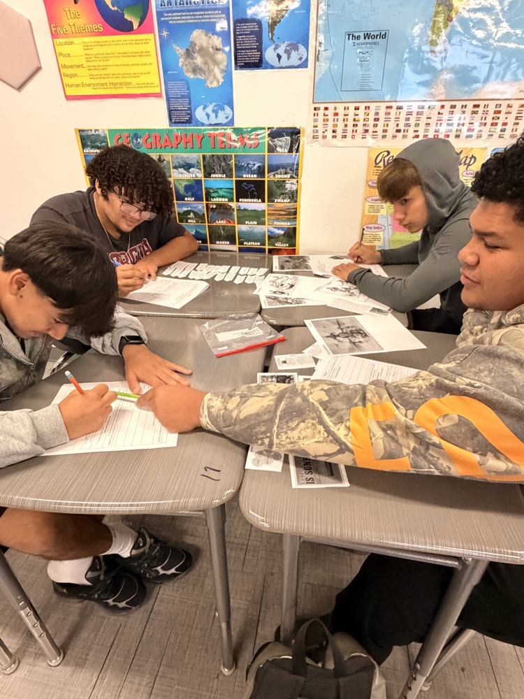 Two students seated at tables, one pointing at a paper with a pen, both 