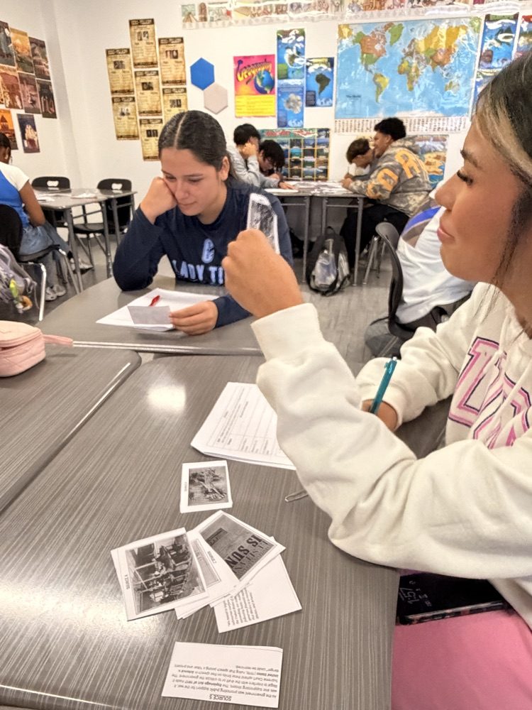 Two students seated at tables, one pointing at a paper with a pen, both 