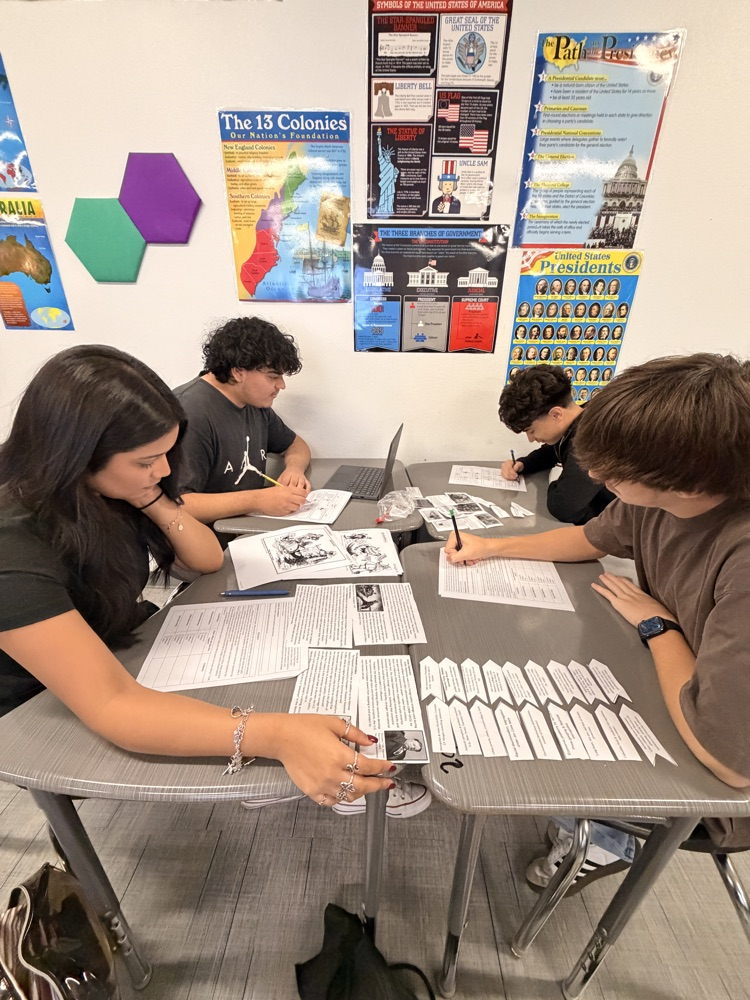 Two students seated at tables, one pointing at a paper with a pen, both 