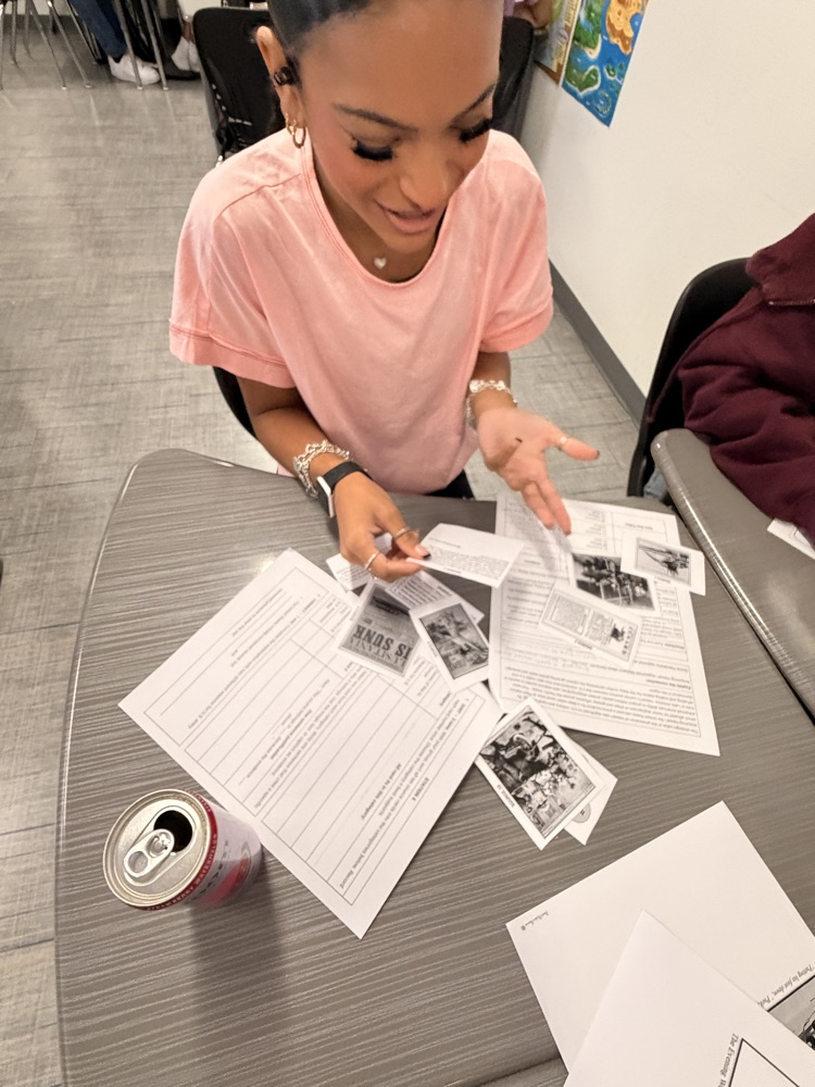A person sitting at a table with papers and a can. The person is wearing a pink shirt.
