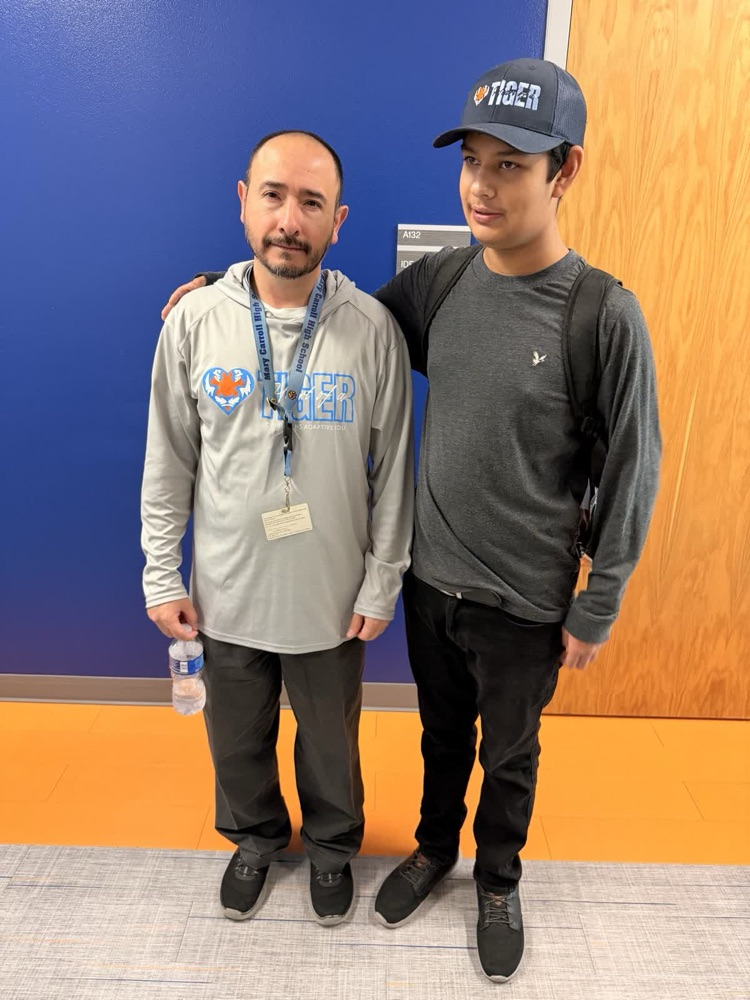 Two men stand in a hallway; one holds a water bottle and wears a cap. Both have IDs, one with a hoodie and the other in a gray shirt.