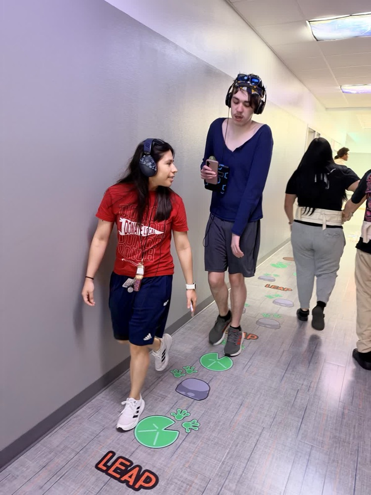 Two men stand in a hallway; one holds a water bottle and wears a cap. Both have IDs, one with a hoodie and the ot