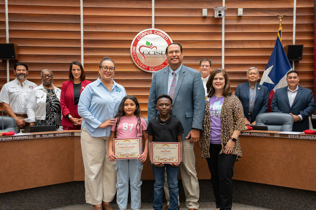 People stand in a room, with two children holding certificates. Behind them is a wall with a logo and a flag.