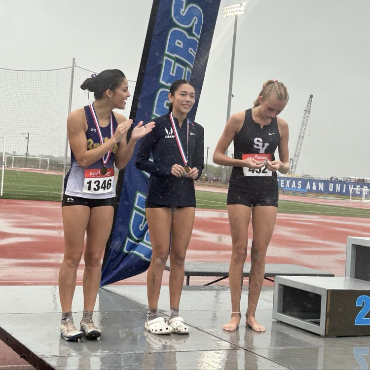 Three athletes on stage, each holding a medal, one with a blue banner behind them.