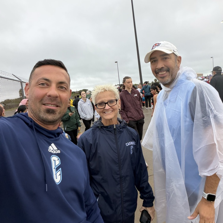 Three people stand outdoors; a man in a blue hoodie, a woman in a blue jacket, and a man in a white poncho.