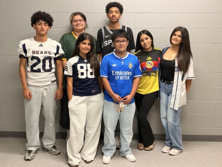 Group of students standing together in front of a wall, each wearing different colored sports jerseys.