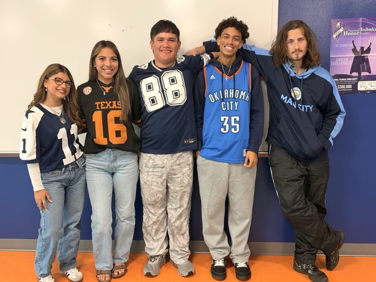 Five students in sports jerseys stand close together against a white wall with an orange floor.