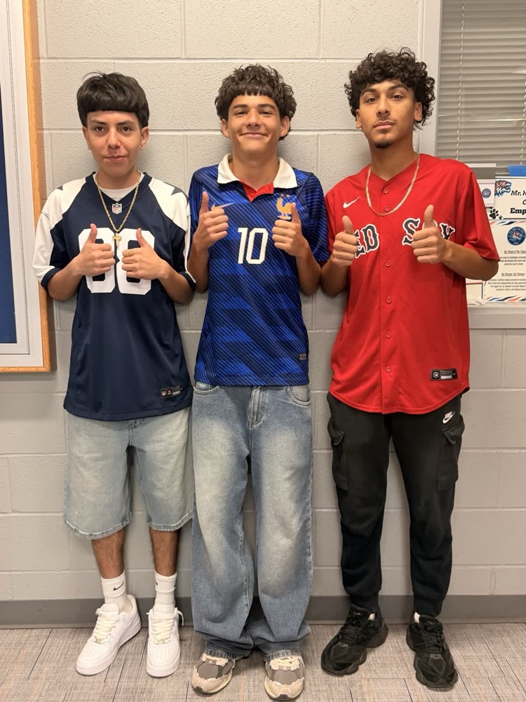 Three boys stand together, smiling. One wears a red shirt, another blue, and the third wears denim.