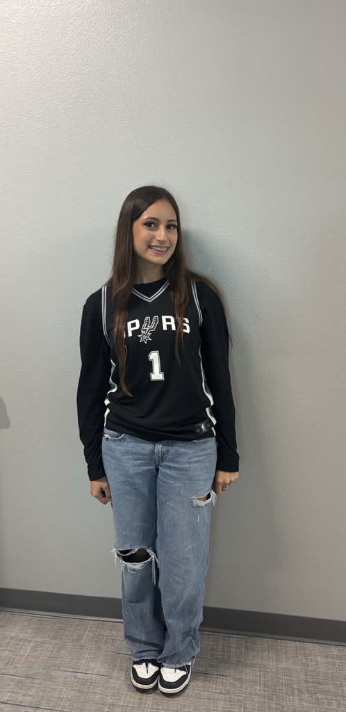 Person in black jersey, number 1, standing against a white wall, wearing jeans and sneakers.
