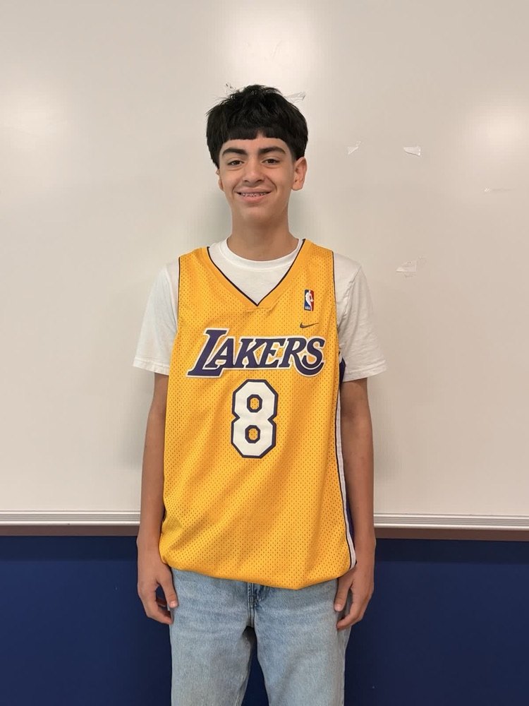 A young boy wearing a Lakers jersey with the number 8 stands in front of a whiteboard.