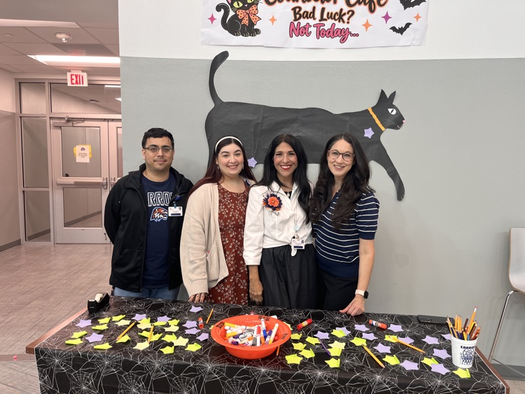 Five people stand behind a table with a Halloween theme, a large black cat on the wall.