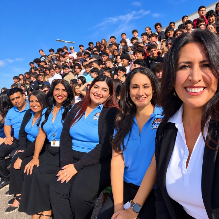 Four women in blue uniforms pose with large crowd seated in stadium bleachers. Bright sky above.