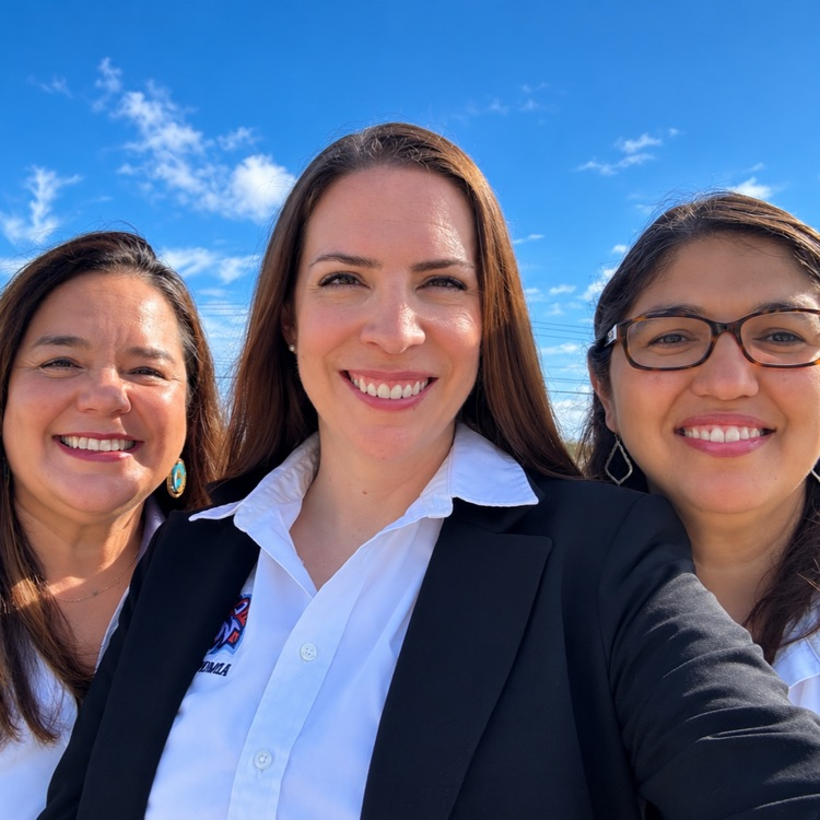 Three women smiling, standing close together in front of a blue sky. The woman in middle wearing a black blazer and white shirt.