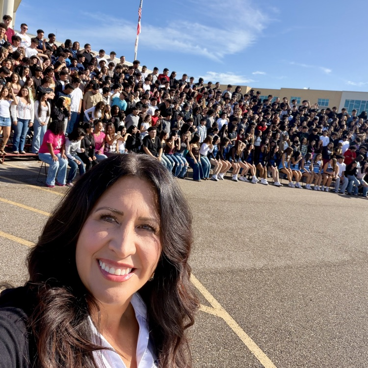 A woman smiling in front of a large crowd sitting on bleachers; a school building is in the background.