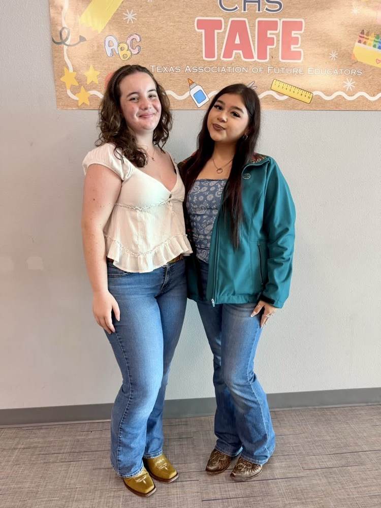 Two students stand close, smiling, against a wall with a banner above them. One wears white blouse, blue jeans, and gold shoes. The other wears a teal jacket and dark blue jeans.
