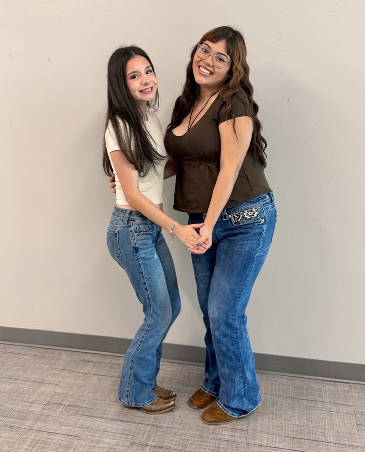 Two girls stand in front of a white wall, holding hands. One wears a white shirt, jeans, and brown boots. The other wears a brown shirt, jeans, and brown boots.