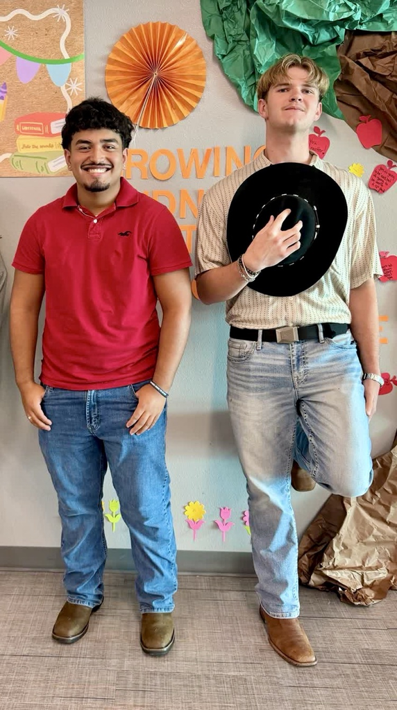 Two young men stand beside each other, one holding a hat. They wear light blue jeans and brown boots. A colorful wall backdrop with a tree and fruit is behind them.