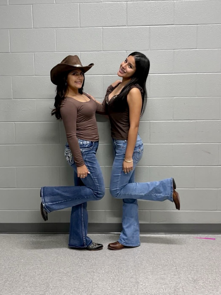 Two women standing, one wearing a cowboy hat, both wearing jeans, standing against a brick wall.