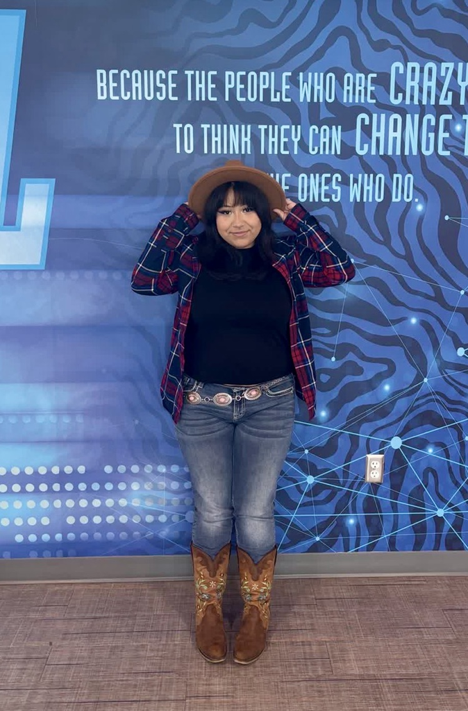 Person standing in front of wall with quote, wearing hat, plaid shirt, jeans, and brown boots.