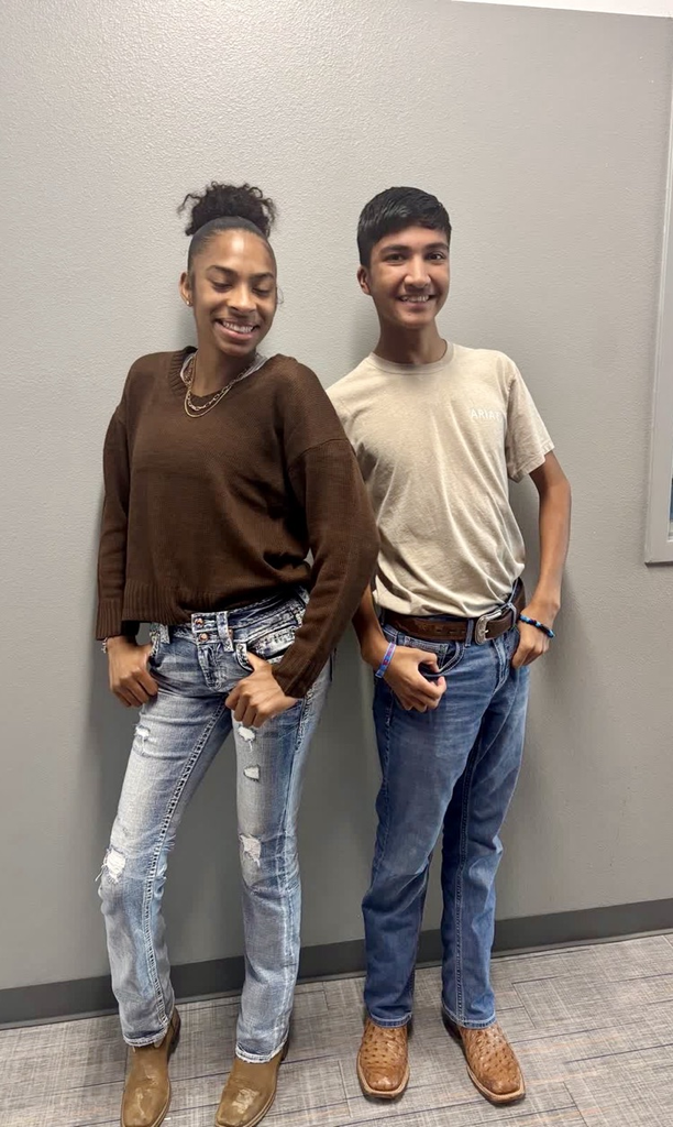 Two students, one in brown top and jeans, the other in beige shirt and jeans, stand next to each other with smiles and hands in pockets.
