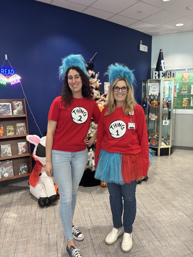 Two women in costumes with blue hair stands in front of a Christmas tree and bookshelf.