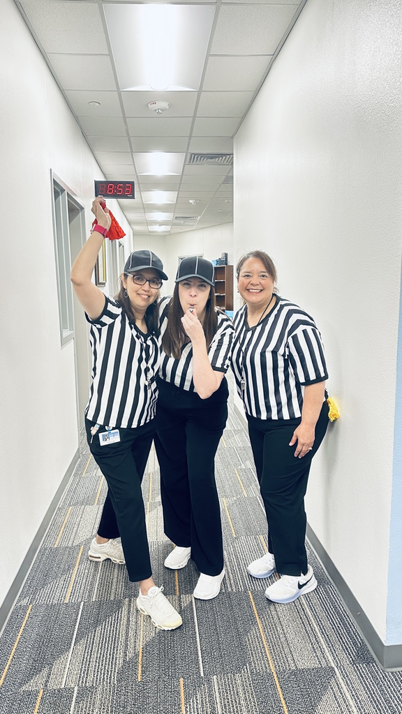 Three women in matching uniforms pose together in a hallway with a digital clock above.