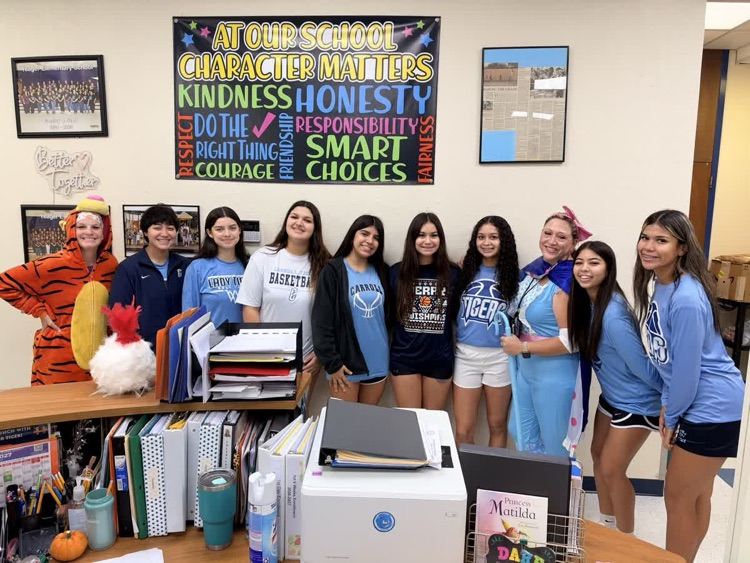 A group of students in matching blue shirts and shorts stand in a classroom, posing for a photo.