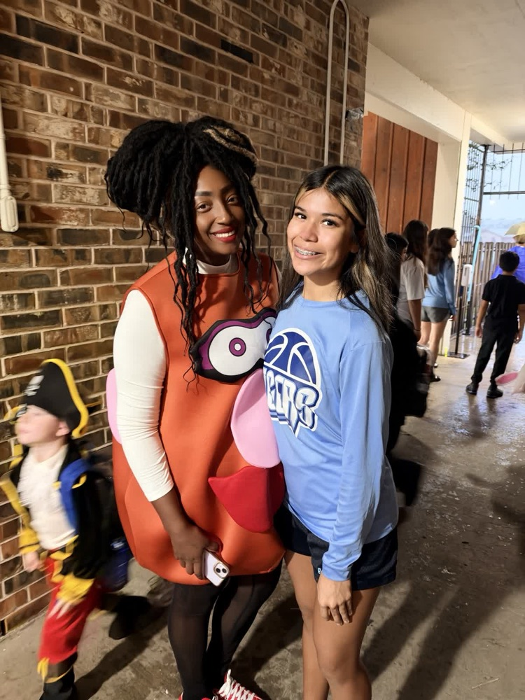 Two girls, one in a costume, smiling for a photo; background shows hallway with brick wall and people.