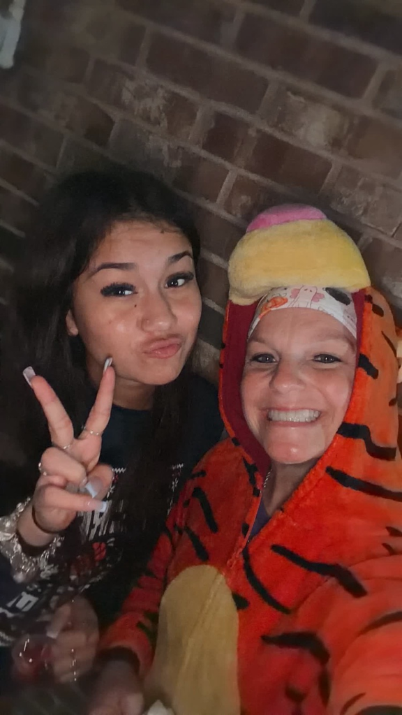 Two girls smile for a photo. One wears a tiger onesie. Behind them is a brick wall.