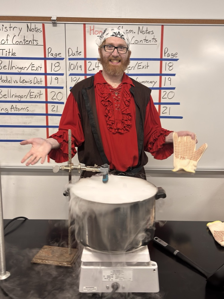 Man in a red shirt with a beard wearing a crown and holding a pot over a hot plate.