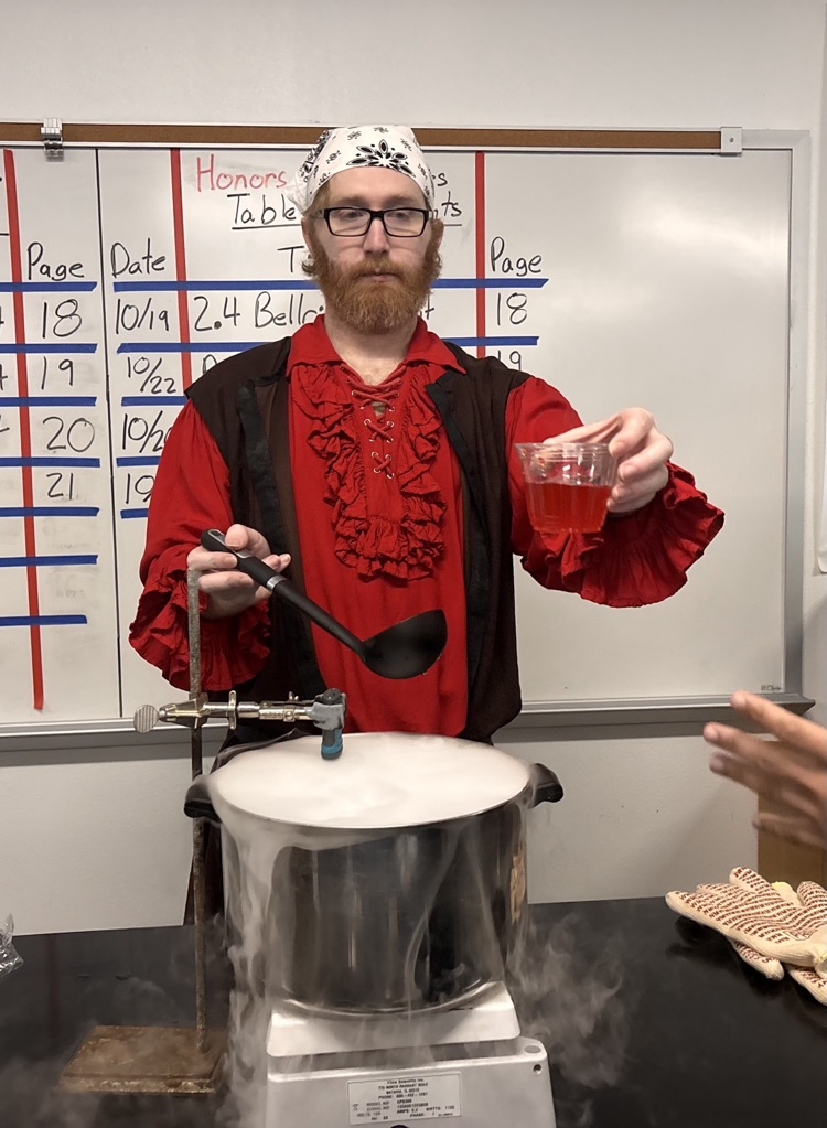 A man holds a ladle and a glass with red liquid in front of a boiling pot.