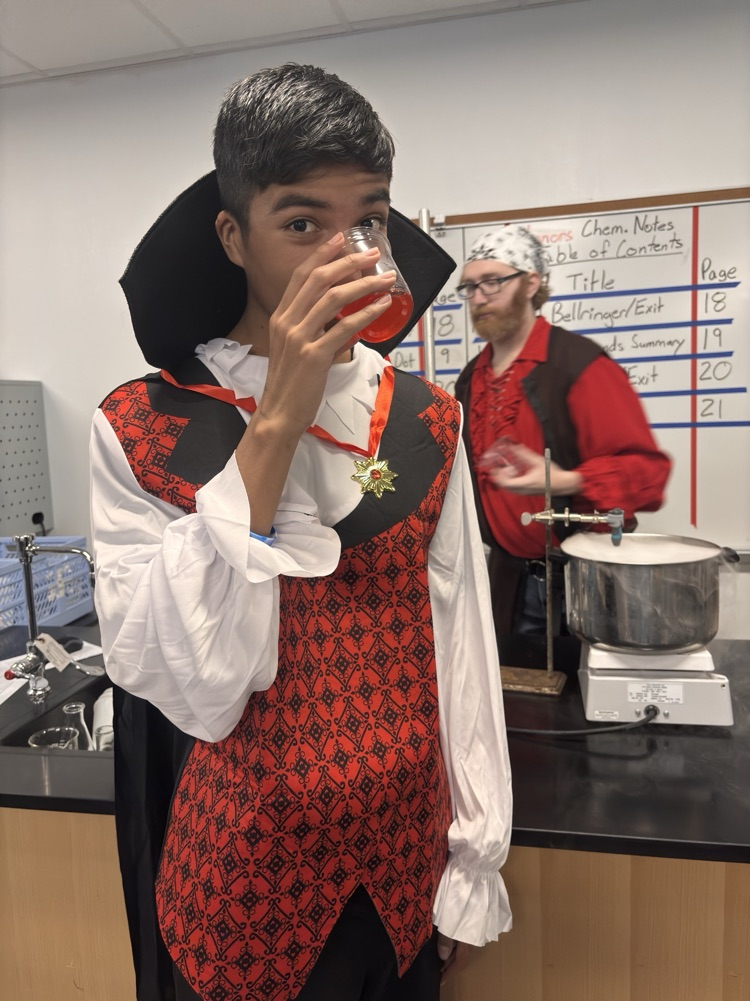A student dressed in a vampire costume drinks a red liquid while standing in a lab.