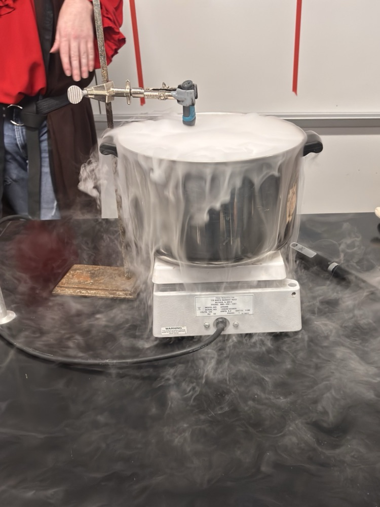 A person in a red long-sleeved shirt standing at a table with a silver pot emitting steam.