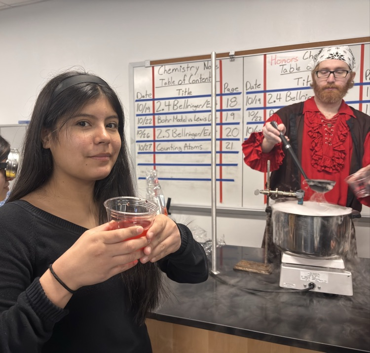 A girl in a black shirt holding a cup of liquid. A person in a red shirt is standing behind a counter holding a pot.