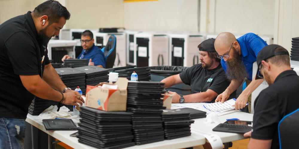 Technology staff sort and stack large batches of laptop computers on a table in a technology workspace.