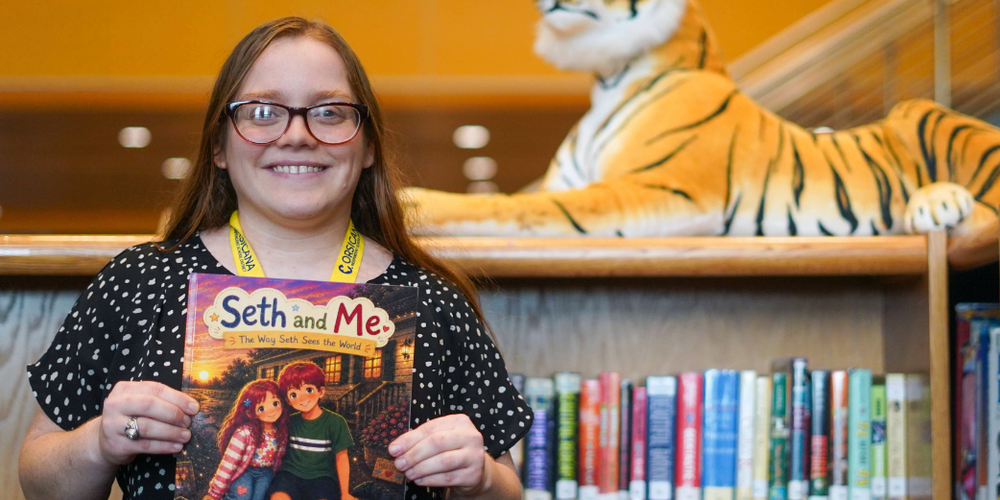Corsicana ISD psychologist Gibson holding a children’s book titled “Seth and Me” in front of a library bookshelf with a large tiger plush  in the background.