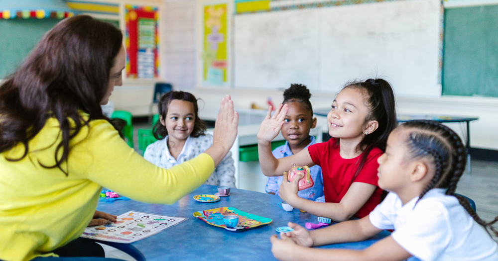 Photograph shows a teacher high-fiving four young students during a classroom activity. 