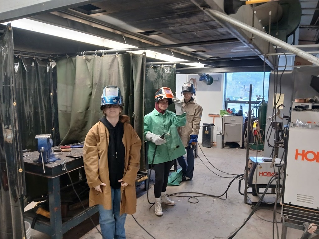 3 young ladies are wearing protective gear and helmets, smiling and getting ready to weld!