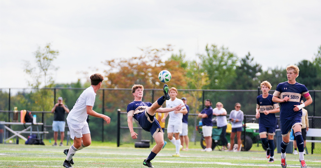 group of soccer plaers with one player kicking the ball with trees in the backround