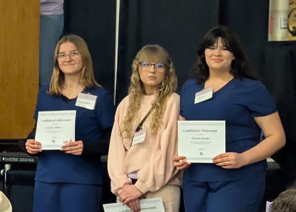Three women in blue uniforms hold certificates. One wears glasses, another wears a light pink top.