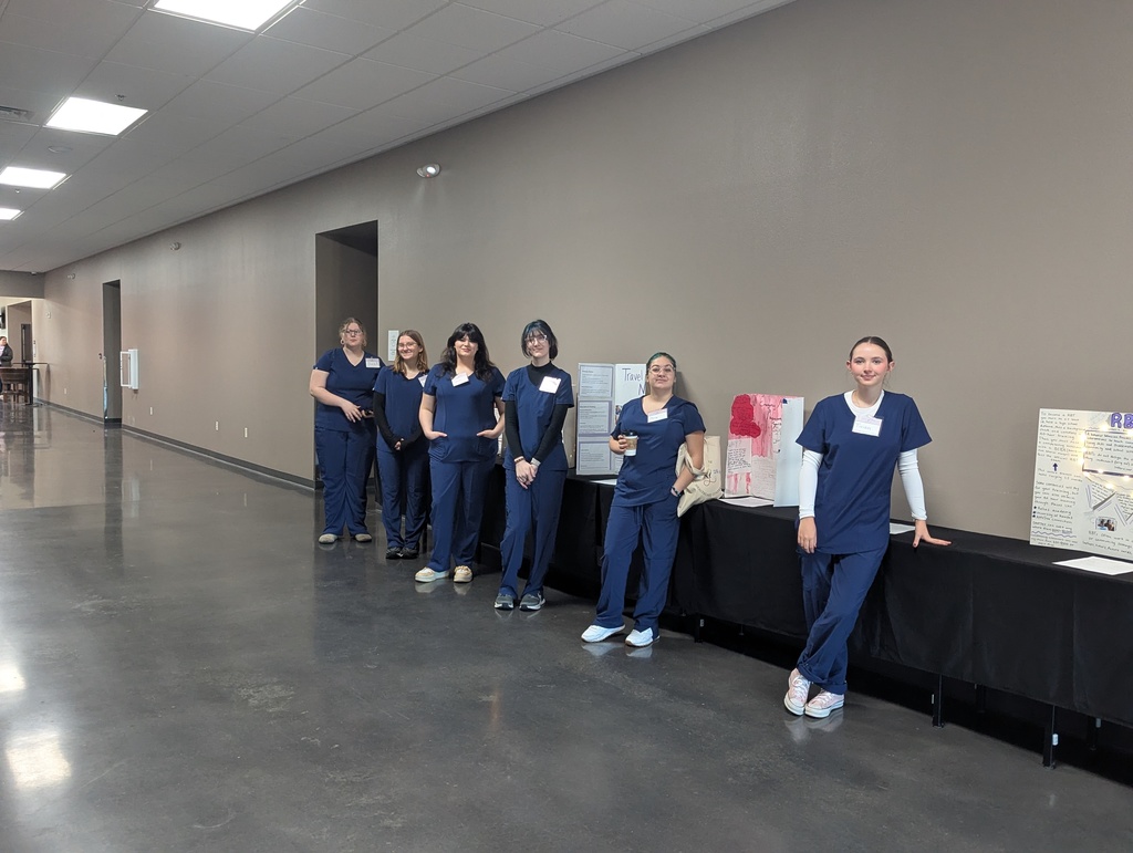 A group of seven women in blue uniforms stand near a table in a corridor with beige walls and ceiling lights.